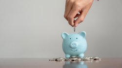 A blue piggy bank on top of a pile of silver coins. A hand puts a silver coin in the piggy bank. 